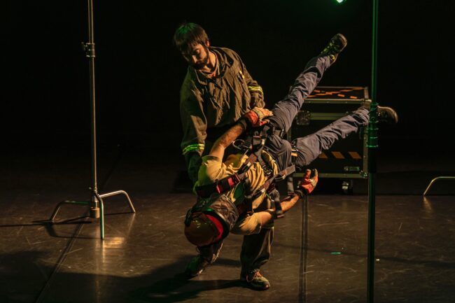 Jon Ander Urresti - Eukalyptus. Plano general, dos figuras masculinas en el escenario, ambas vestidas de bombero. Una de las figuras en pie, sostiene a la otra, estática, girada 60 grados en el aire.