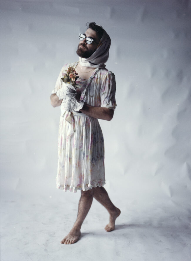 Jon Ander Urresti - Foto de estudio. Prueba de vestuario, atuendo femenino, vestido con motivo floral, descalzo. Cabeza medio cubierta con un pañuelo, guantes blancos, sostiene un ramo de flores.