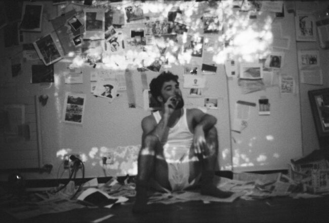 Jon Ander Urresti - Retrato plano medio. Blanco y negro. Instantánea tomada en el estudio. Sentado en el suelo contra la pared, fumando. La pared está cubierta de fotografías, notas de prensa y bocetos.