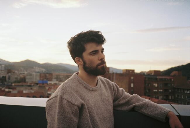 Jon Ander Urresti - Retrato plano medio. En la azotea de un edificio, al fondo se ven otros bloques de viviendas, y montañas. Cielo abierto a primera hora de la mañana.